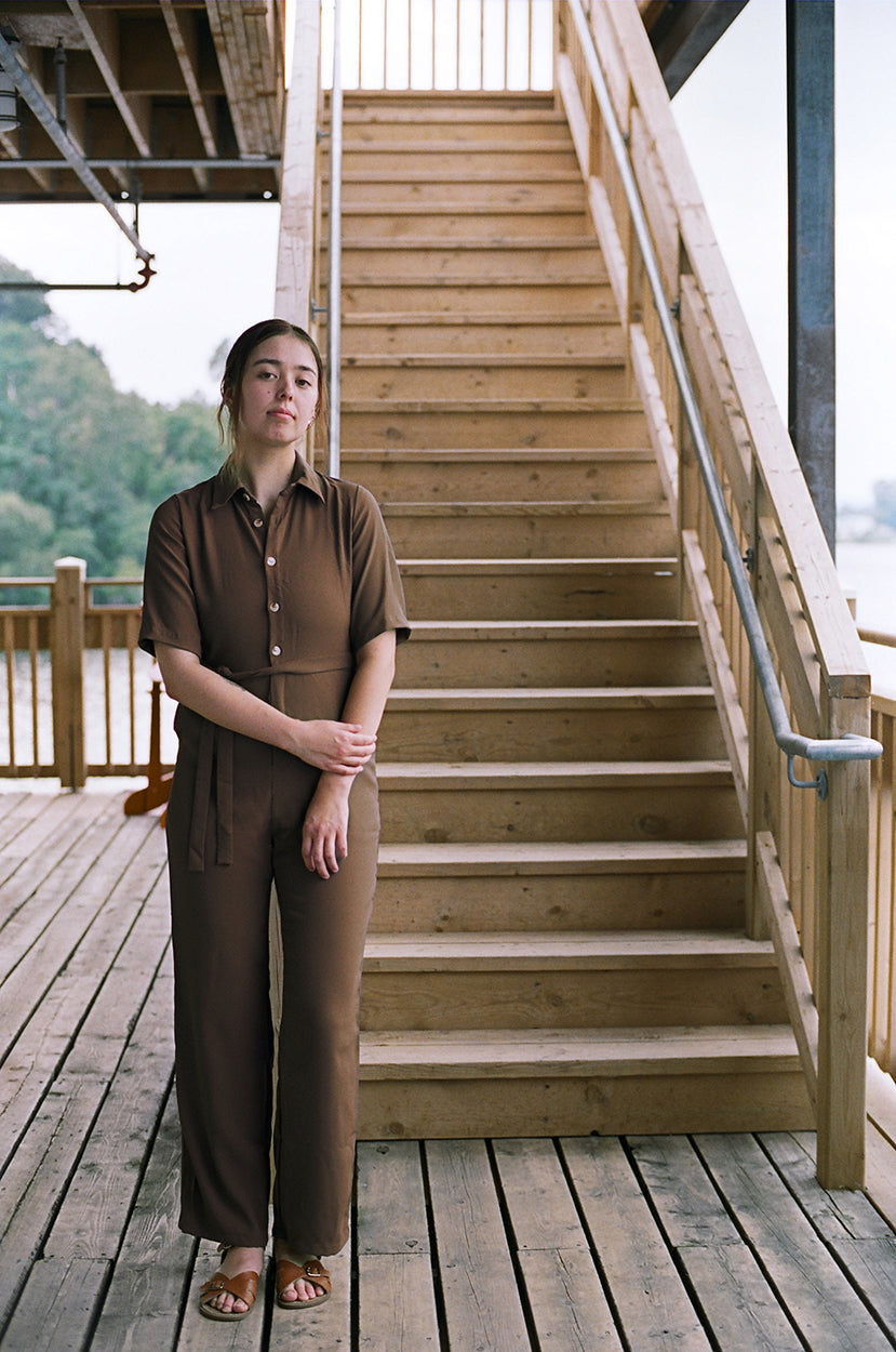 Model standing up, leaning against the a staircase, looking at the camera, brown coloured jumpsuit, brazilian crepe patou,