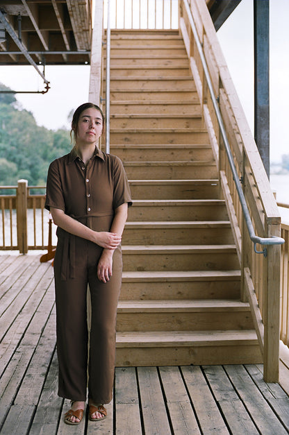 Model standing up, leaning against the a staircase, looking at the camera, brown coloured jumpsuit, brazilian crepe patou,